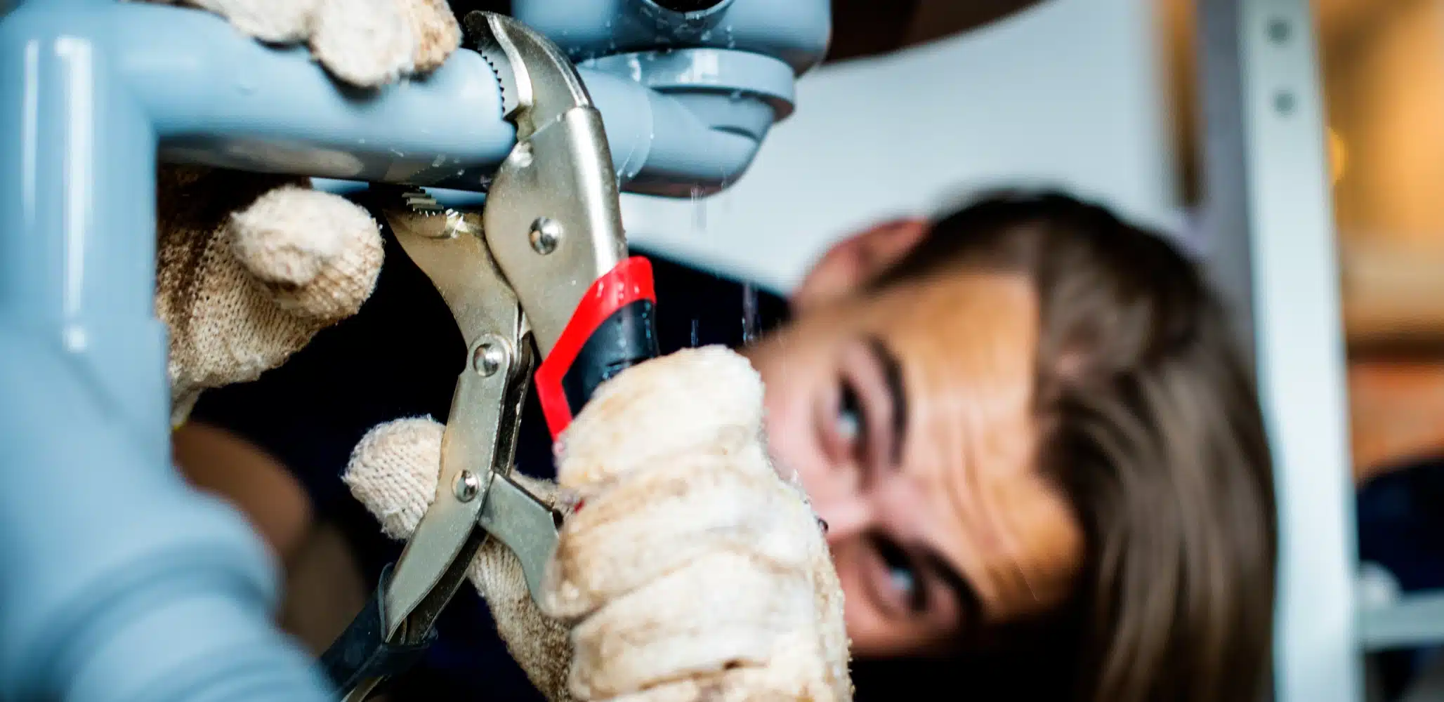 A plumbing expert wearing gloves uses pliers to fix a leaking gray pipe under a sink. Water droplets are visible near the pipe connection, showcasing the careful attention to detail provided by heating experts in Salt Lake City.