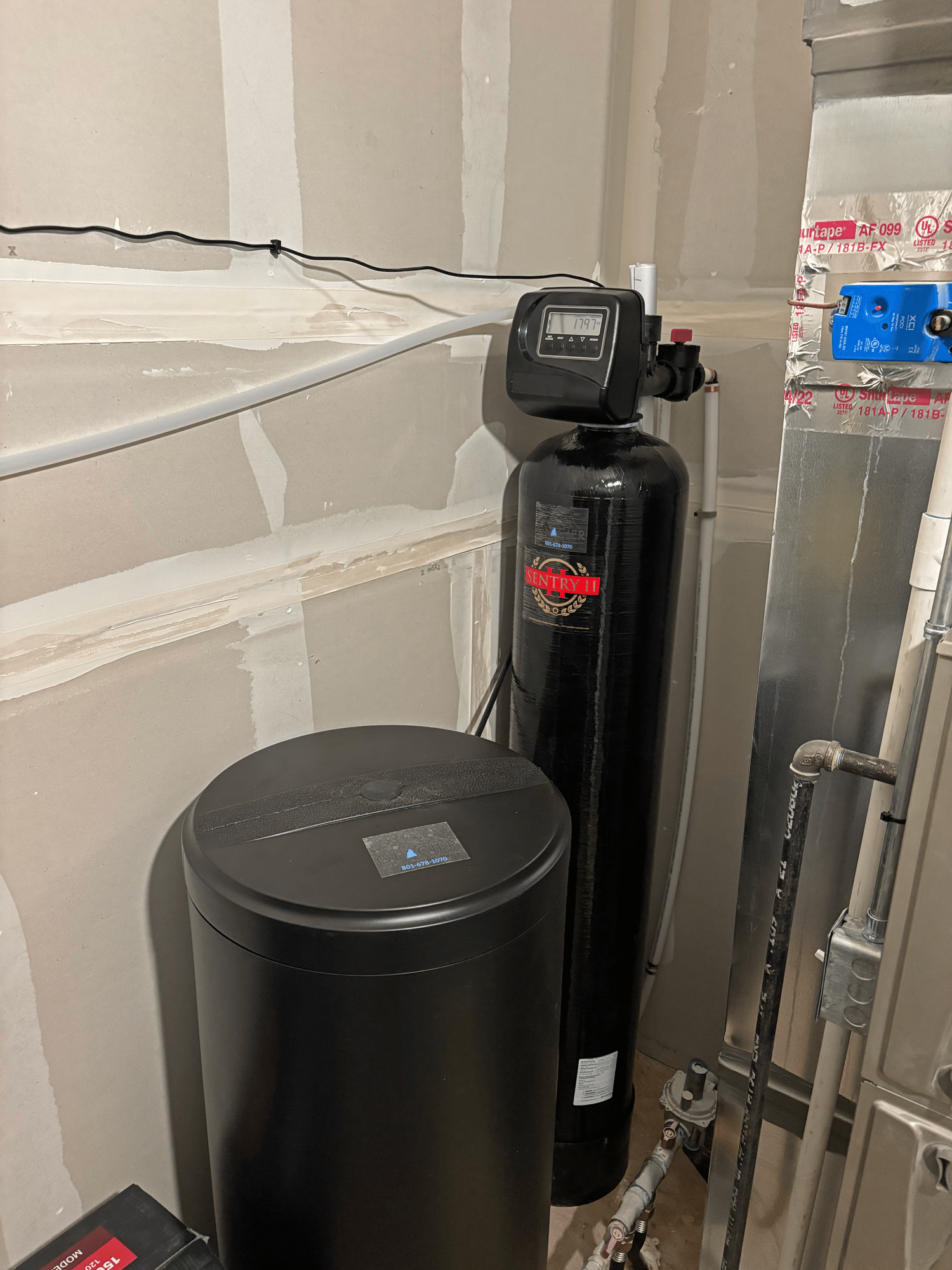 A water softener system is installed in a basement corner with exposed drywall and pipes visible nearby, highlighting the professional touch of Plumbing and Heating Experts in Salt Lake City.