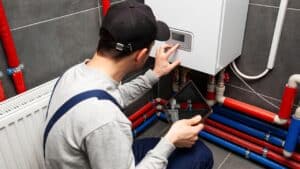 A technician in a cap and uniform uses a tablet while adjusting settings on a wall-mounted water heater with visible red and blue pipes, highlighting the tank vs. tankless options available in Utah homes.