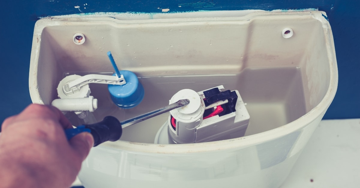 Hand using a screwdriver to adjust or repair the inside components of a toilet tank, with visible fill valve and flush mechanism - a typical task for Plumbing & Heating Experts in Salt Lake City.