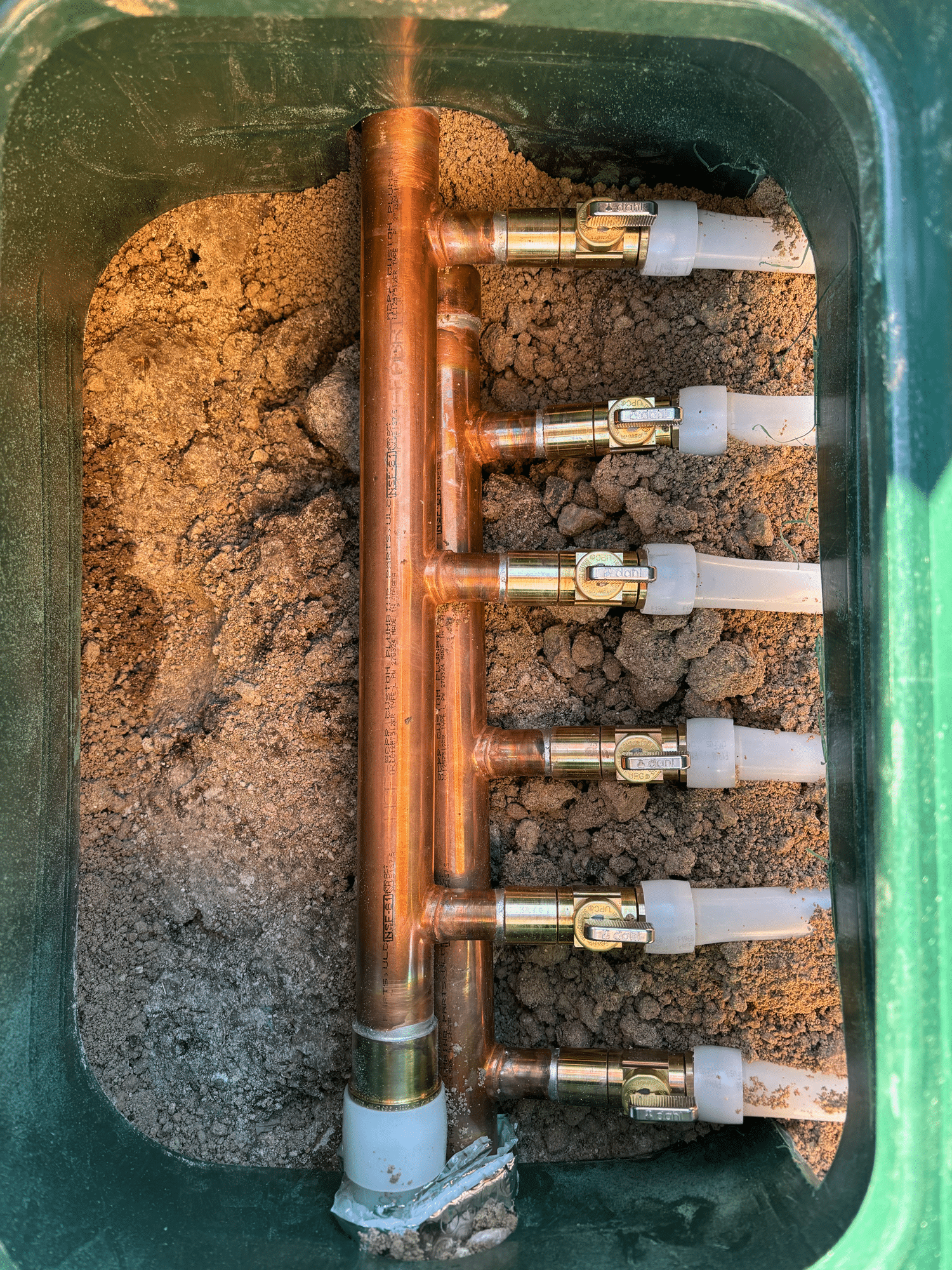 A copper water manifold with six brass valves and white pipe connections, expertly installed by plumbing and heating experts in Salt Lake City, sits securely in a green utility box surrounded by soil.