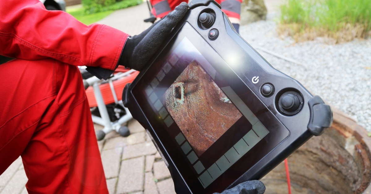 A person in red overalls and black gloves holds a rugged tablet displaying a close-up image of a rusted metal object, outdoors near gravel and a manhole - showcasing the careful inspection skills of plumbing and heating experts in Salt Lake City.