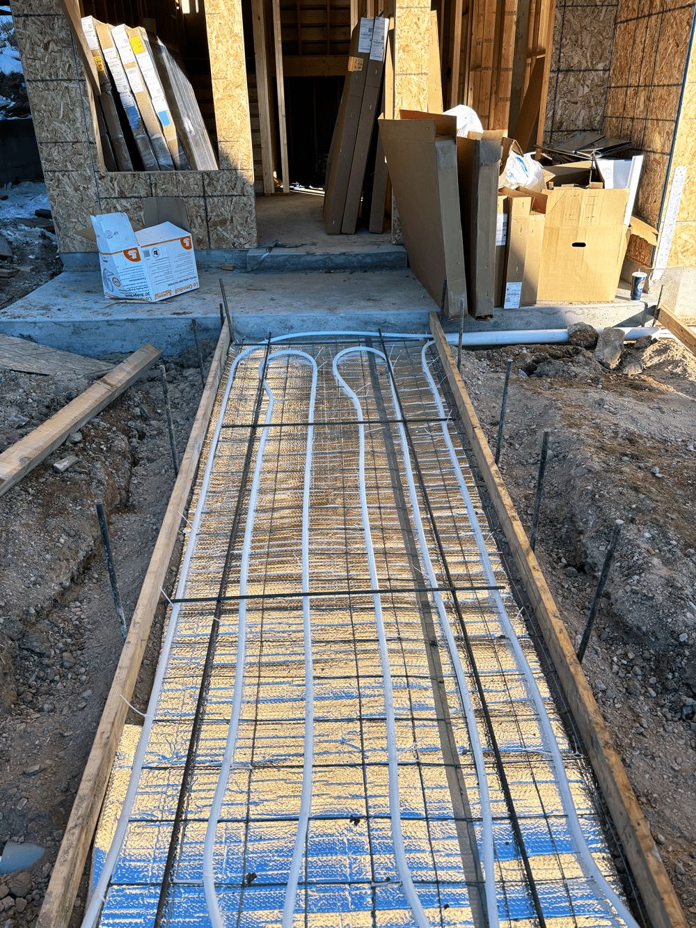 White tubing for a radiant heating system, expertly installed by heating experts in Salt Lake City, runs through a concrete walkway mold at a construction site; building materials are stacked near a framed house in the background.