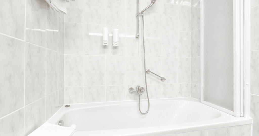 A white bathtub with a wall-mounted showerhead, two soap dispensers, a grab bar, and a frosted shower screen creates a comfortable and functional space in this tiled bathroom - showcasing thoughtful plumbing solutions often recommended by heating experts in Salt Lake City.