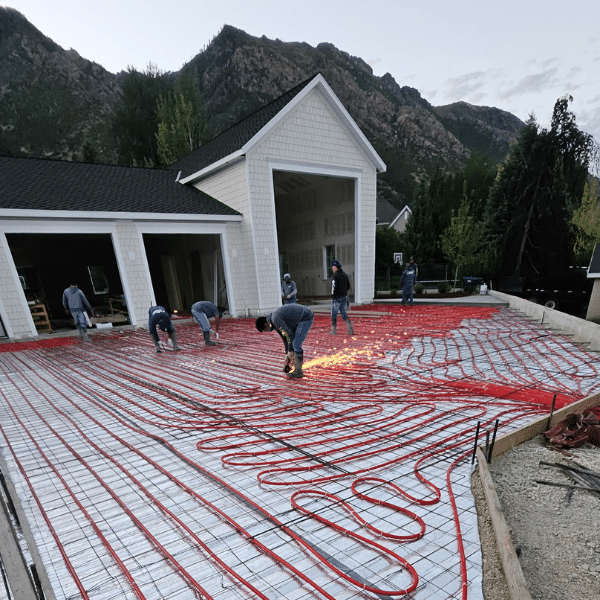 Workers install red radiant heating pipes in a grid pattern across a driveway in front of a large white garage with mountains in the background, demonstrating how heated driveways work and prompting the question: are heated driveways worth it?.