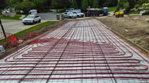 Red tubing for heated driveways is arranged in a grid pattern on a driveway, with construction vehicles and equipment in the background—giving insight into installation and heated driveway cost.