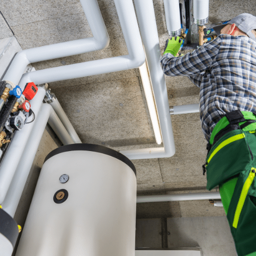 A worker in protective gloves and uniform installs or repairs insulated pipes connected to a large tankless water heater system in a utility room.