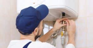 Person wearing a blue cap and white shirt adjusting pipes beneath a wall-mounted water heater in a tiled room, showcasing the skill of plumbing and the expertise of heating experts in Salt Lake City.