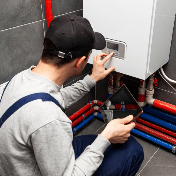 A person in work clothes inspects and adjusts the settings on a wall-mounted water heater during a boiler installation, holding a digital tablet near red and blue pipes.