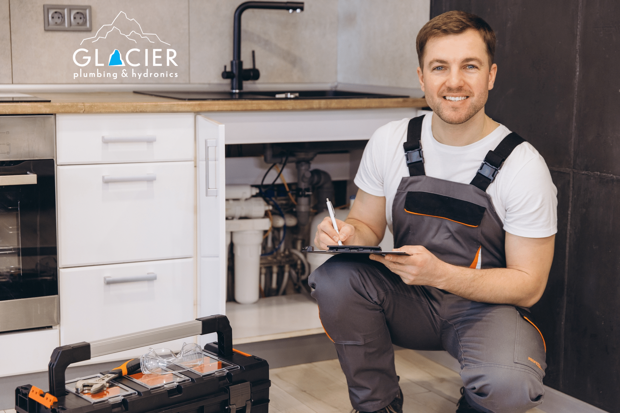 A Salt Lake City plumber in work overalls kneels by an open kitchen sink cabinet, holding a clipboard and pen, with a toolbox nearby. The Glacier Plumbing & Hydronics logo is visible, highlighting trusted home plumbing services in Utah, based out of Salt Lake City. Leak repair for water leaks, heater and boiler repair, water heater flush services, it appears Glacier Plumbing does it all for residents.