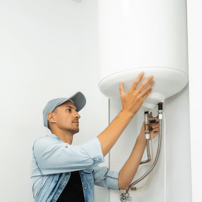A man wearing a cap inspects or performs water heater repair on a white unit mounted on the bathroom wall.