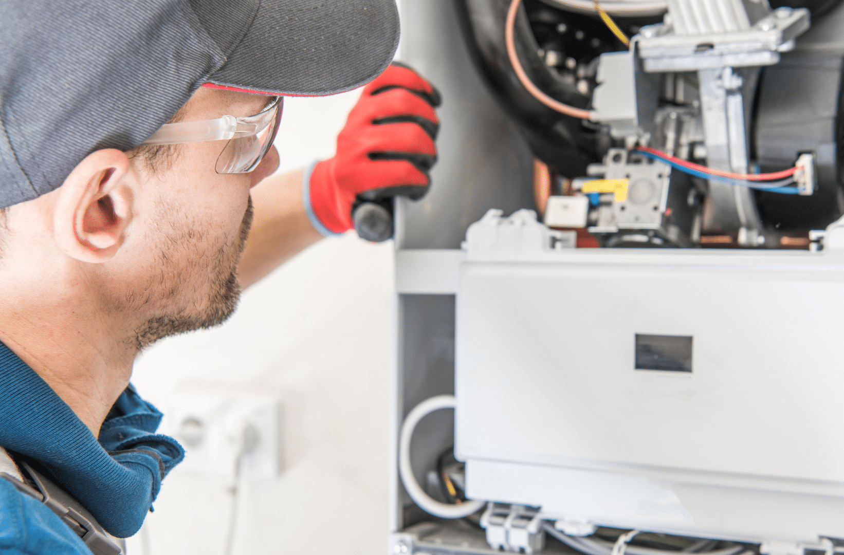A technician wearing safety glasses and red gloves inspects the interior of a gas furnace, holding a flashlight near electrical components - just one example of the thorough attention to detail provided by Heating Experts in Salt Lake City, who specialize in both heating and plumbing services.