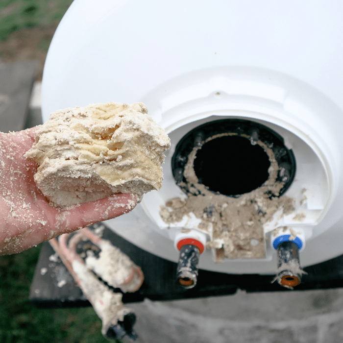 A hand holds a chunk of limescale sediment removed from the open bottom of a water heater, with mineral buildup visible around the access point—a clear sign that water heater repair may be needed.