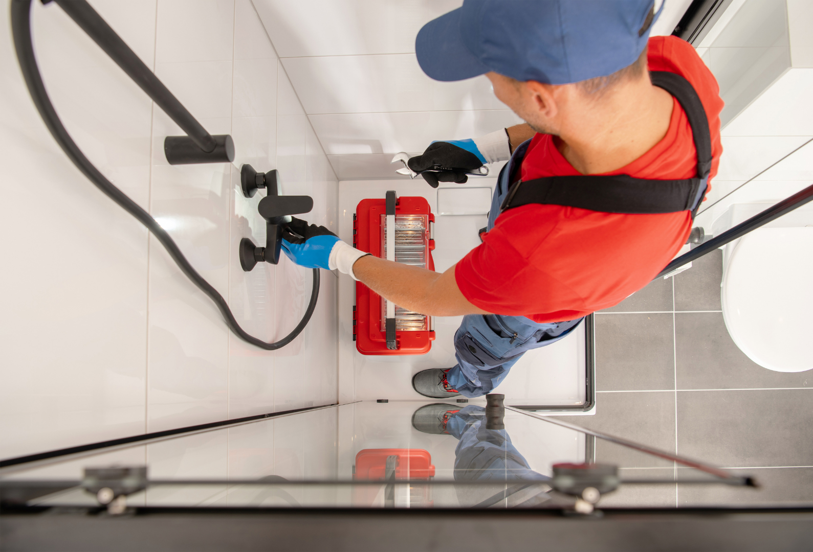 A worker in gloves and overalls installs or repairs a shower faucet in a tiled bathroom using tools from a red toolbox, demonstrating the skill and precision you can expect from Plumbing and Heating Experts in Salt Lake City.