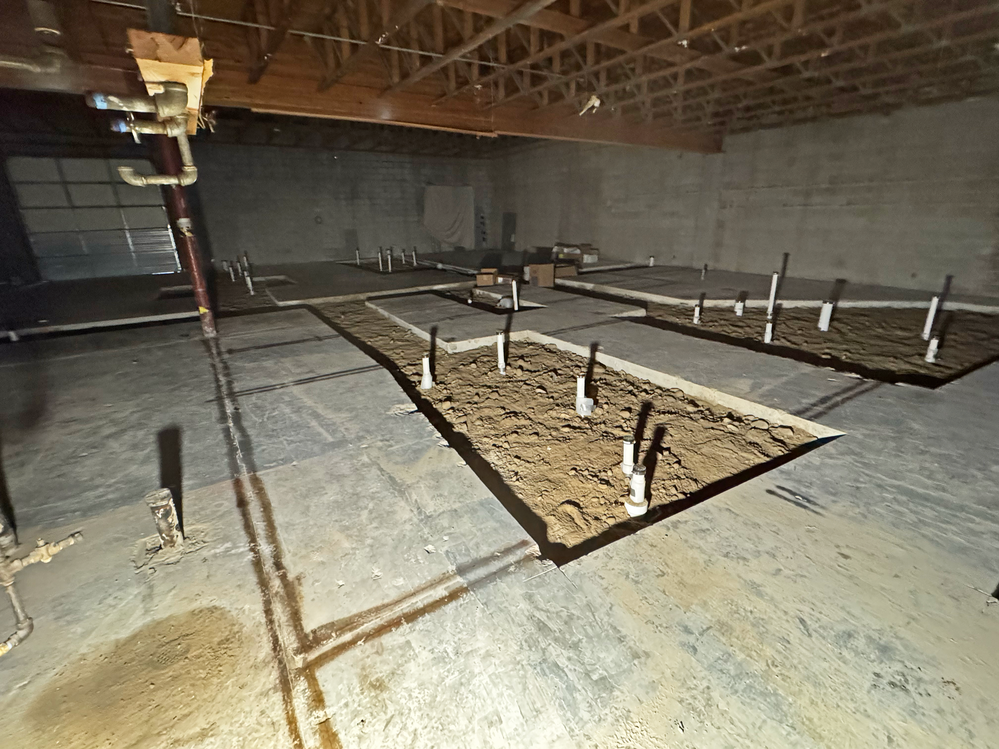 Large, unfinished indoor construction site with exposed dirt sections, plumbing pipes sticking up, and visible ceiling beams - ideal for Plumbing and Heating Experts in Salt Lake City to work their magic.