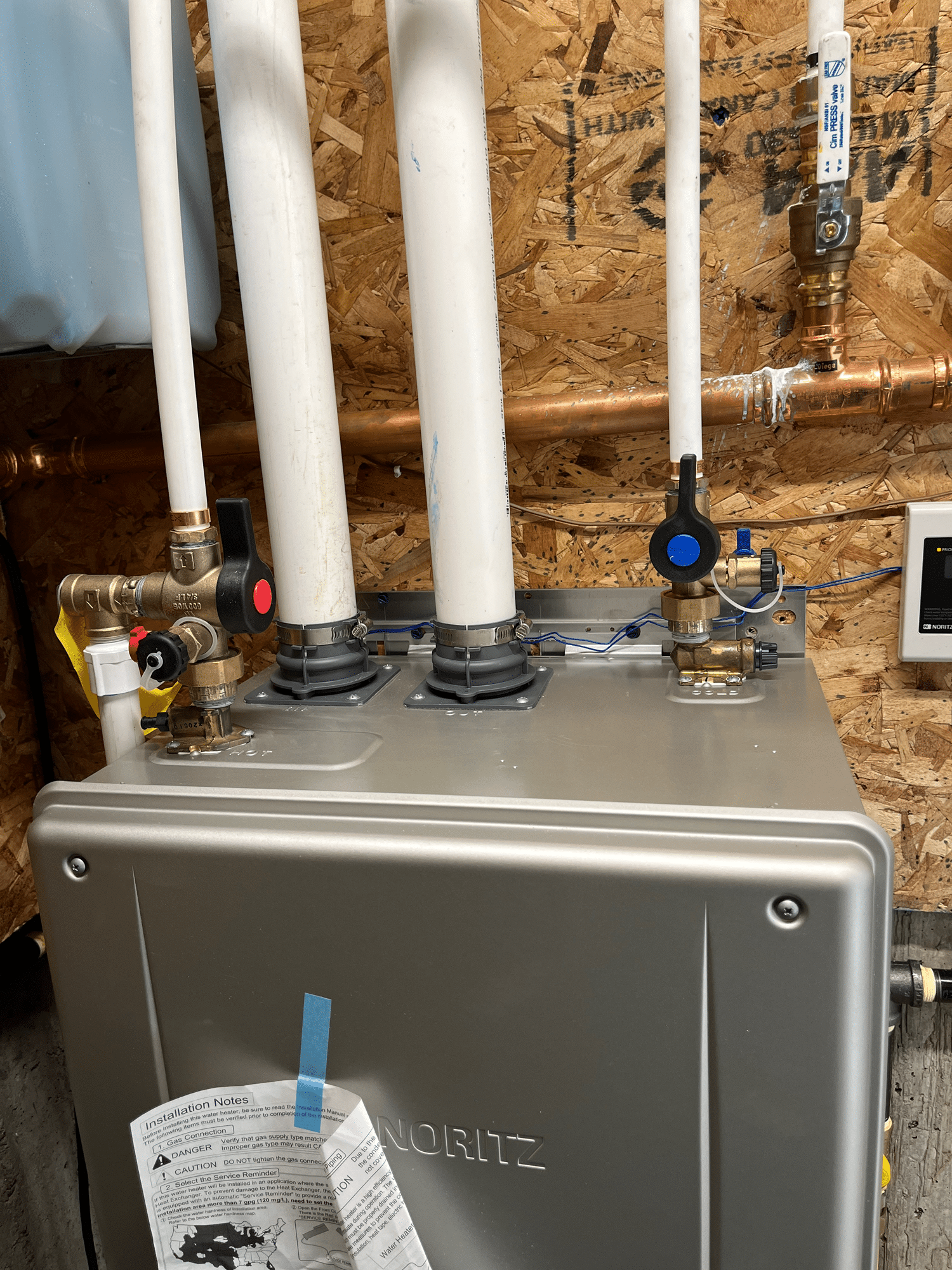 Tankless water heater installed on an unfinished wall, with labeled PVC pipes, metal connectors, and an installation manual taped to the front - a testament to the precision and care provided by heating experts in Salt Lake City specializing in plumbing solutions.