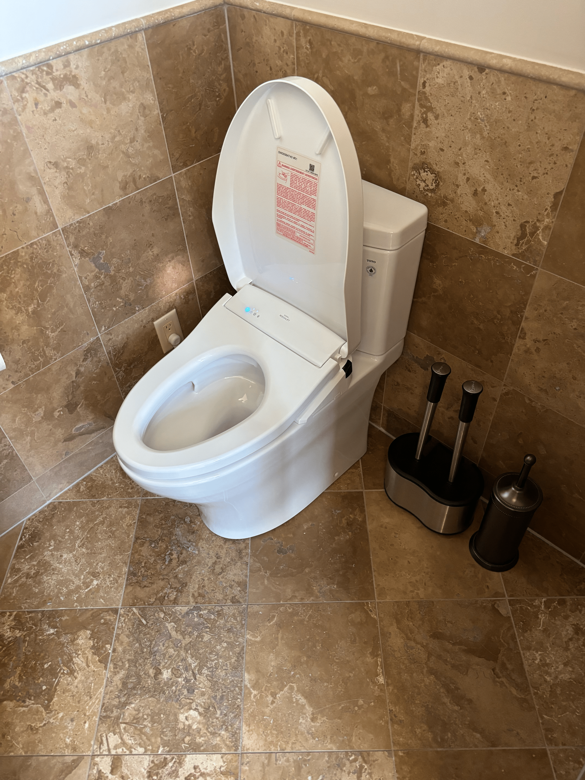Modern white toilet with lid open, set against tan tiled walls and floor. Two toilet brushes and a metal trash can are placed beside it, showcasing the neat attention to detail you’d expect from local plumbing and Heating Experts in Salt Lake City.