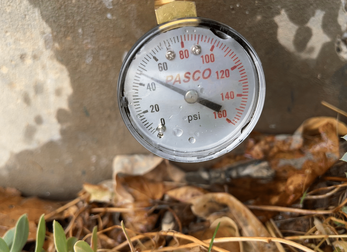 A close-up of a PASCO pressure gauge outdoors, reading approximately 22 psi, with water droplets on the glass and dry leaves in the background - an everyday scene for Heating Experts in Salt Lake City handling plumbing systems in changing weather conditions.